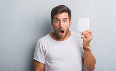 Gri grunge duvar holding boş defter bir sürpriz yüz, korkuyor ve heyecanlı korku ifadesi ile şok korkmuş yakışıklı genç adam