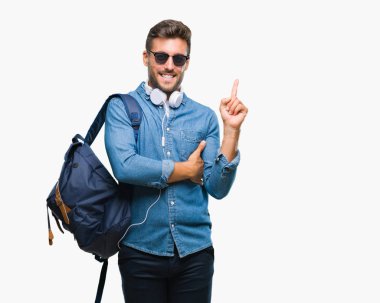 Yüzünde kocaman bir gülümseme ile izole arka plan üzerinde kulaklık ve sırt çantası giyen genç yakışıklı turist adam kameraya bakarak yan el ve parmak ile işaret.