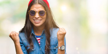 Genç güzel Arap kadın üzerinde güneş gözlüğü takıyor arka plan çok mutlu ve heyecanlı kaldırdı, silah ile kazanan jest yapıyor gülümseyen ve başarı için çığlık izole. Kutlama kavramı.