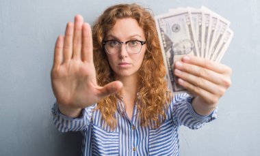 Gri grunge duvar açık eliyle dur işareti ile ciddi ve kendine güvenen ifade, savunma jest yapıyor dolar tutan genç Kızıl saçlı kadın