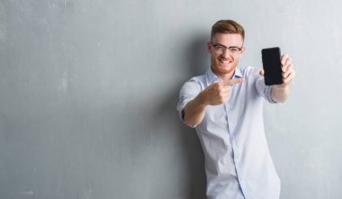 Genç Kızıl saçlı adam gri grunge duvar gösteren boş ekran smartphone el ve parmak ile işaret çok mutlu