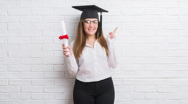 年轻的成年妇女在白色砖墙戴研究生帽持有程度惊讶与一个想法或问题指向手指与幸福的脸 — 图库照片