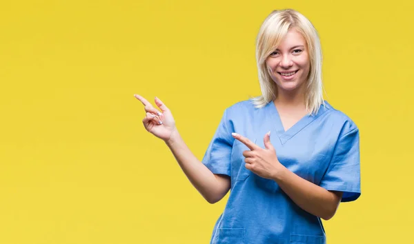 年轻美丽的金发护士医生妇女在孤立的背景微笑 看着相机指着两个手和手指到一边 — 图库照片