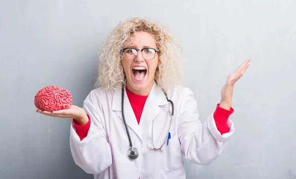 Young blonde doctor woman over grunge grey wall holding brain very happy and excited, winner expression celebrating victory screaming with big smile and raised hands