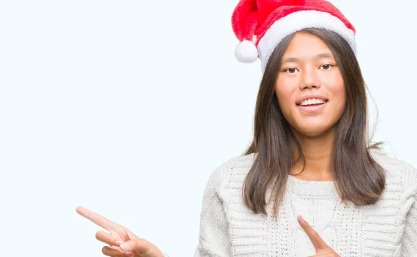 年轻的亚洲妇女戴着圣诞节帽子在孤立的背景微笑 看着相机指向用两个手和手指到一边 — 图库照片