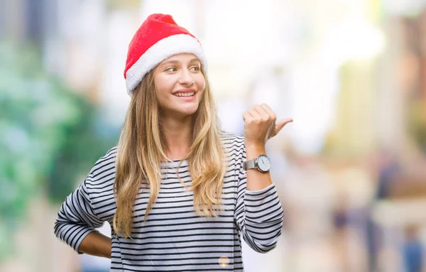 年轻美丽的金发女子戴着圣诞节帽子在孤立的背景微笑与快乐的脸看和指向侧与拇指向上 — 图库照片