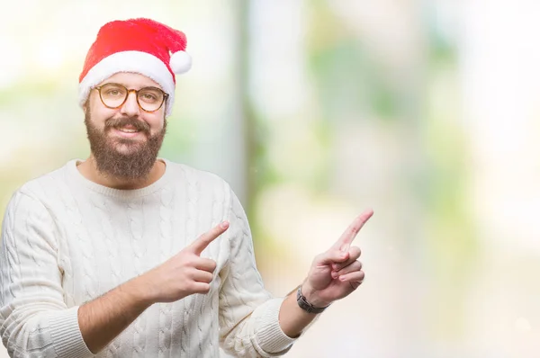 年轻的高加索男子戴着圣诞帽 戴着眼镜在孤立的背景上微笑着看着相机 用两只手和手指指着一边 — 图库照片