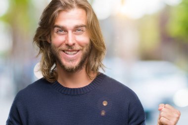 Uzun saçlı izole arka plan üzerinde kutluyor kış kazak giyen genç yakışıklı adam şaşırttı ve başarı için kaldırdı kollarını açarak hayret ve gözleri açık. Kazanan kavramı.