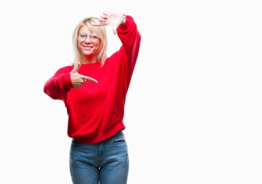 Kazak ve gözlük elle yapma çerçeve ve parmakları ile mutlu yüz gülümseyen izole arka plan üzerinde giyen genç güzel sarışın kadın. Yaratıcılık ve Fotoğrafçılık kavramı.