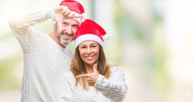 Orta yaş İspanyol çift elle yapma çerçeve ve parmakları ile mutlu yüz gülümseyen izole arka plan üzerinde Noel şapka giyiyor. Yaratıcılık ve Fotoğrafçılık kavramı.