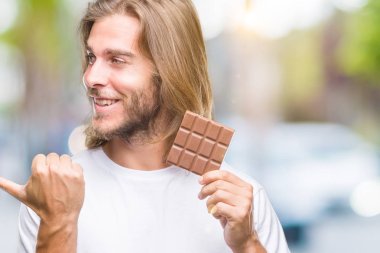 Uzun saç beslenme ile yakışıklı delikanlı çikolata bar işaret ve başparmak kadar mutlu yüz gülümseyen tarafı ile gösteren izole arka plan üzerinde