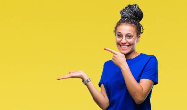 Saç Afro-Amerikan Kız hayran ve el ile sunulması ve parmak ile işaret ederken kameraya gülümseyen izole arka plan üzerinde gözlük takan genç örgülü.