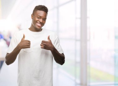 El ile pozitif jest yapıyor beyaz t-shirt başarı işareti giyen genç Afro-Amerikan adam başparmak gülümseyen ve mutlu. Neşeli bir ifadeyle, kazanan jest kameraya bakıyor.