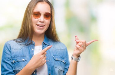 Genç güzel beyaz kadın gülümseyerek ve iki elleri ve parmakları ile tarafına işaret kamera bakarak izole arka plan üzerinde güneş gözlüğü takıyor.