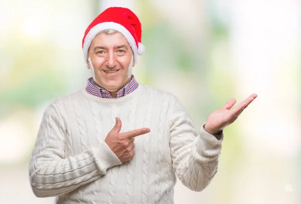 驚くし 手とを提示 指で指している間カメラに笑顔孤立の背景にクリスマス帽子をかぶっているハンサムな年配の男性 — ストック写真