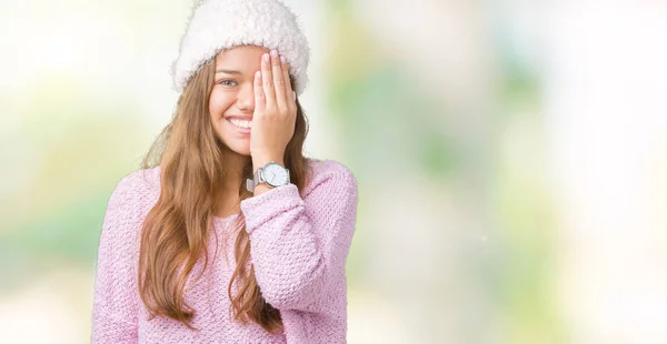 顔と驚きの感情に自信を持って笑顔で手で片目を覆う孤立の背景にセーターを着て 冬の帽子を着て若い美しいブルネットの女性 — ストック写真
