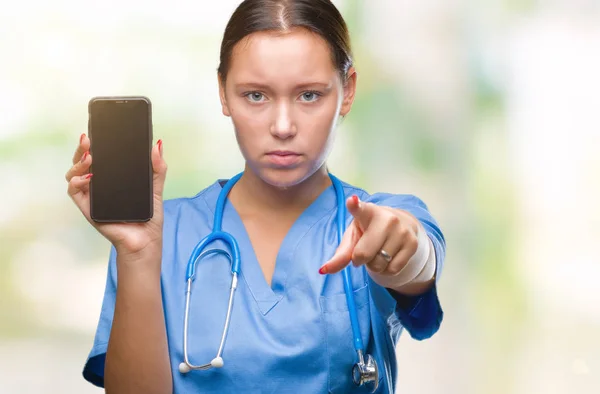 年轻的白种人医生妇女显示 Smarpthone 在孤立的背景指向与手指相机和你 正面和自信的手势从前面 — 图库照片