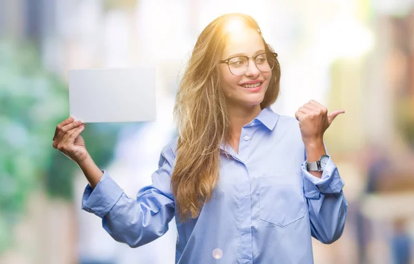 Young beautiful blonde business woman holding blank card over isolated background pointing and showing with thumb up to the side with happy face smiling