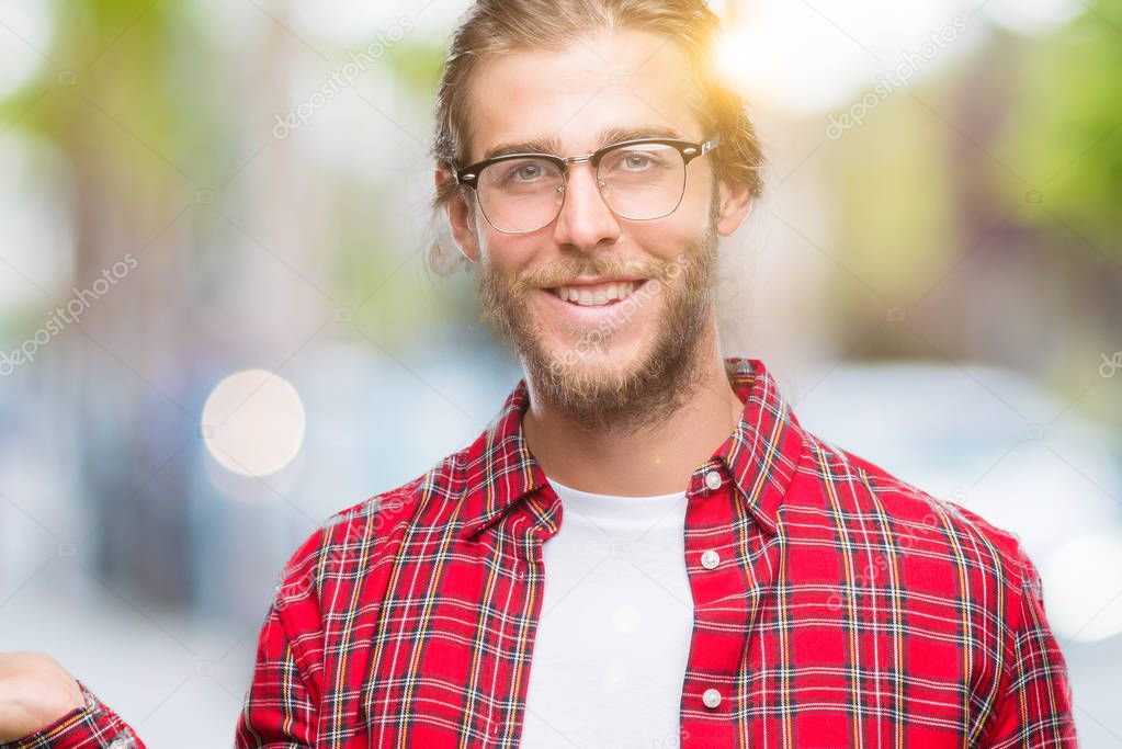 Young handsome man with long hair wearing glasses over isolated background smiling cheerful presenting and pointing with palm of hand looking at the camera.