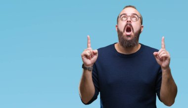 Güneş gözlüğü izole arka plan üzerinde giyen genç beyaz hipster adam hayret ve yukarıya bakıp parmakları ile işaret şaşırttı ve silah kaldırdı.