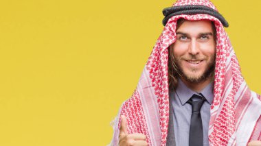 Genç yakışıklı Arap adam uzun saçlı keffiyeh izole arka plan başarı işareti el ile pozitif jest yapıyor üzerinde giyen başparmak gülümseyen ve mutlu. Neşeli bir ifadeyle, kazanan jest kameraya bakıyor.