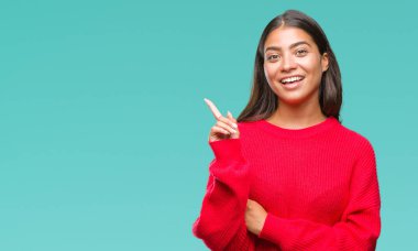 Kameraya bakarak yan el ve parmak ile işaret yüzünde kocaman bir gülümseme ile izole arka plan üzerinde kış kazak giyen genç güzel Arap kadın.