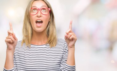 Hayret ve yukarıya bakıyor ve parmakları ile işaret ve silah kaldırdı sürpriz izole arka plan üzerinde gözlüklü genç güzel kadın.