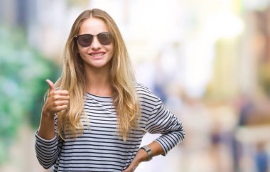 Genç güzel sarışın kadın hareketi el ile mutlu yaşasın yapıyor izole arka plan üzerinde güneş gözlüğü takıyor. Kameraya bakarak başarı gösteren ile ifade onaylama.