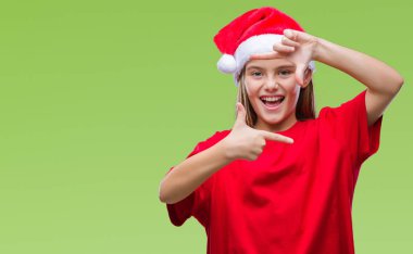Genç güzel kız elle yapma çerçeve ve parmakları ile mutlu yüz gülümseyen izole arka plan üzerinde Noel şapka giyiyor. Yaratıcılık ve Fotoğrafçılık kavramı.