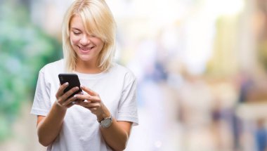 Ayakta ve diş gösterilen kendine güvenen bir gülümsemeyle gülümseyerek mutlu bir yüz ile izole arka plan üzerinde akıllı telefon kullanan genç güzel sarışın kadın