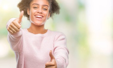 El ile pozitif jest yapıyor onaylama izole arka plan üzerinde kış kazak giyen genç afro Amerikan kadın başparmak gülümseyen ve başarı için mutlu. Kamera, kazanan jest arıyorsunuz.