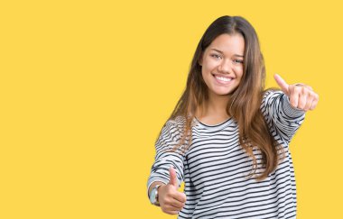 El ile pozitif jest yapıyor onaylama izole arka plan üzerinde çizgili süveter giyen genç güzel esmer kadın başparmak gülümseyen ve başarı için mutlu. Kamera, kazanan jest arıyorsunuz.