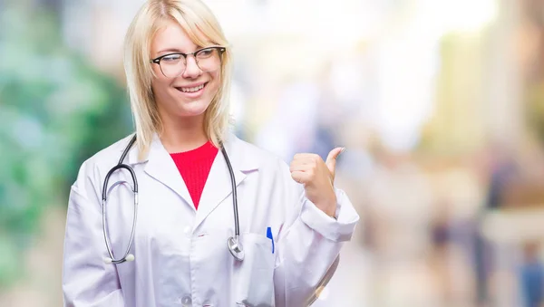 Young beautiful blonde doctor woman wearing medical uniform over isolated background smiling with happy face looking and pointing to the side with thumb up.