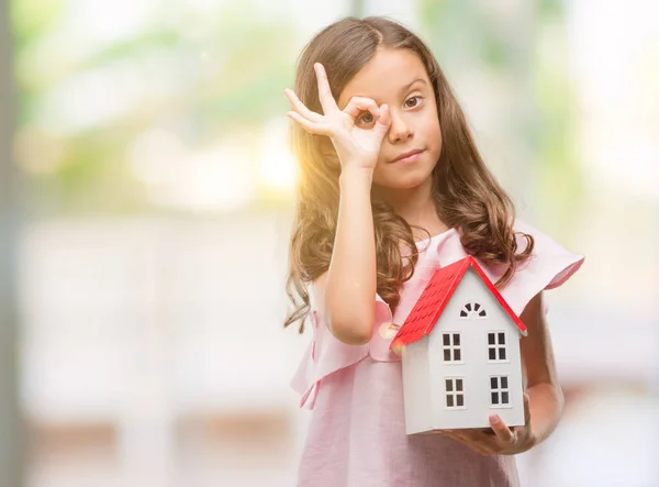 指の間から見て目に手で サインをして笑っている幸せそうな顔で小さな家を保持しているヒスパニックのブルネットの少女 — ストック写真