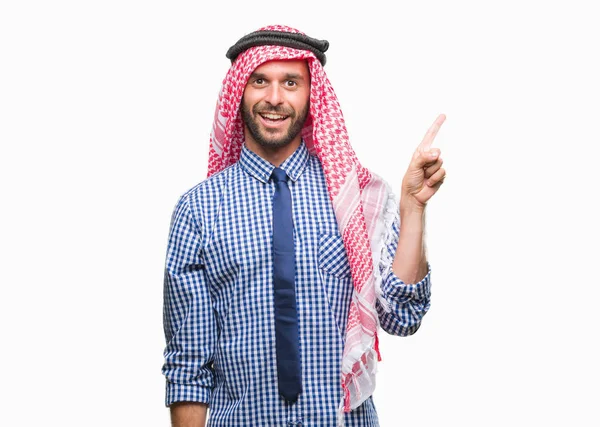 若いハンサムなアラビアン ビジネス男の顔に大きな笑みを浮かべて隔離された背景にクーフィーヤを着てカメラを見て側の手と指で指す — ストック写真