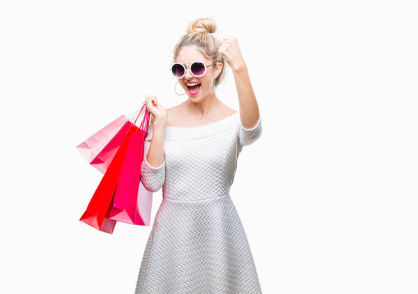 Young beautiful blonde woman holding shopping bags over isolated background annoyed and frustrated shouting with anger, crazy and yelling with raised hand, anger concept