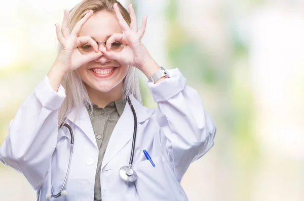 Young blonde doctor woman over isolated background doing ok gesture like binoculars sticking tongue out, eyes looking through fingers. Crazy expression.