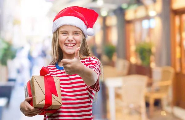 クリスマスの帽子を身に着けている美しい少女 サインをしている笑顔に満足して孤立の背景の上持株ギフトは優秀な兆候の指で親指 — ストック写真