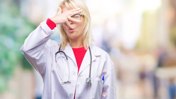美しい金髪女医女性顔と手で目を覆うショックでピーク分離背景上医療ユニフォームを着て顔に指を通して見る — ストック写真