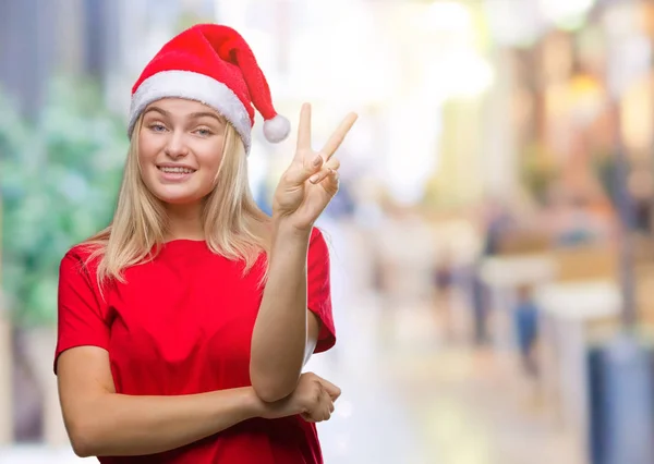 若い白人女性の勝利のサインを行うカメラでウィンクしている幸せそうな顔を浮かべて隔離された背景にクリスマス帽子をかぶっています — ストック写真