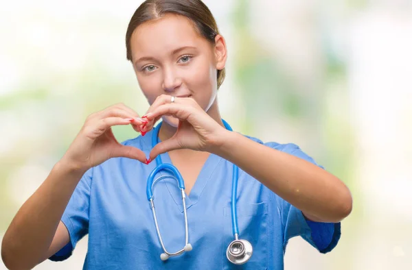 Young caucasian doctor woman wearing medical uniform over isolated background smiling in love showing heart symbol and shape with hands. Romantic concept.