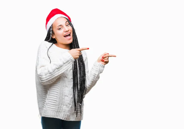 年轻的编织头发非洲裔美国女孩戴着圣诞节帽在孤立的背景微笑着 看着相机指向镜头 用两只手和手指指着一边 — 图库照片