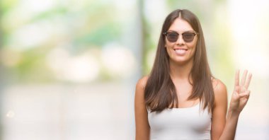 Kendinden emin ve mutlu gülümseyerek sırasında gösterilen ve parmakları ile işaret genç güzel İspanyol giyen güneş gözlüğü üçüncüsü.