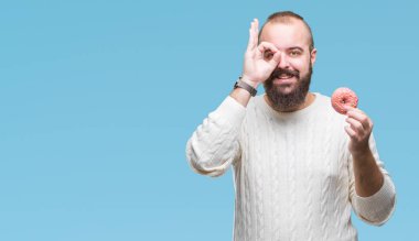 Genç beyaz hipster man ile mutlu yüz el parmaklarının arasından seyir göz üzerinde ok işaretiyle yapıyor gülümseyen izole arka plan üzerinde tatlı çörek yemek