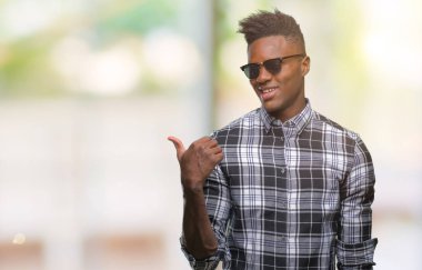Afro-Amerikan delikanlı arıyor ve başparmak yanında yukarıyı mutlu yüz ile gülümseyen izole arka plan üzerinde güneş gözlüğü takıyor.