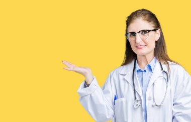 Orta yaş olgun doktor kadın neşeli sunulması ve kameraya bakarak avucunun ile işaret gülümseyen izole arka plan üzerinde tıbbi bir palto giyiyor.