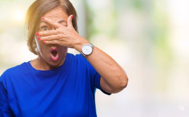 Mahcup ifadeyle parmaklarımın arasından seyir orta yaş İspanyol kadın kıdemli yüz ve gözler el ile kapsayan şok bakmak izole arka plan üzerinde.