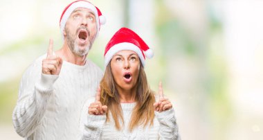 Orta yaş İspanyol çift izole arka plan üzerinde Noel şapka hayret ve yukarıya bakıp parmakları ile işaret şaşırttı ve silah kaldırdı.