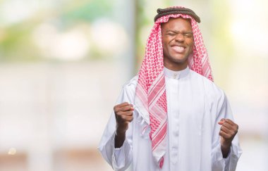 Geleneksel keffiyeh üzerinde giyen genç Arapça Afrika adam arka plan çok mutlu ve heyecanlı kaldırdı, silah ile kazanan jest yapıyor gülümseyen ve başarı için çığlık izole. Kutlama kavramı.