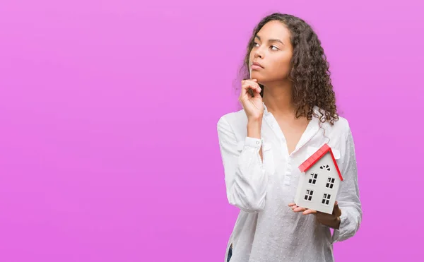Young hispanic real state agent woman holding small house serious face thinking about question, very confused idea
